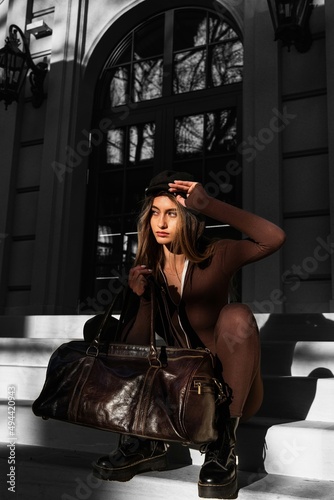 Fashionable young brunette woman with long legs wearing solid long sleeve bodycon one piece jumpsuits posing with a leather brown travel bag