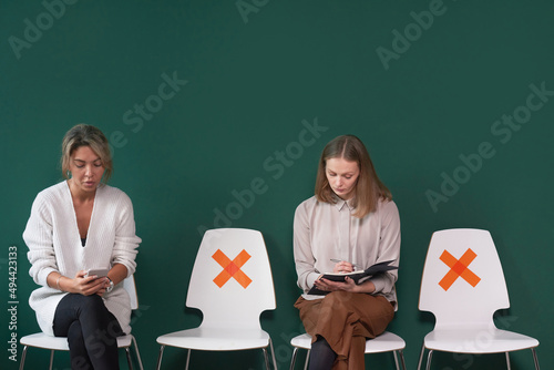 Two elegant young adult women sitting on chairs in queue for job interview against tidewater green wall background, social distance concept photo