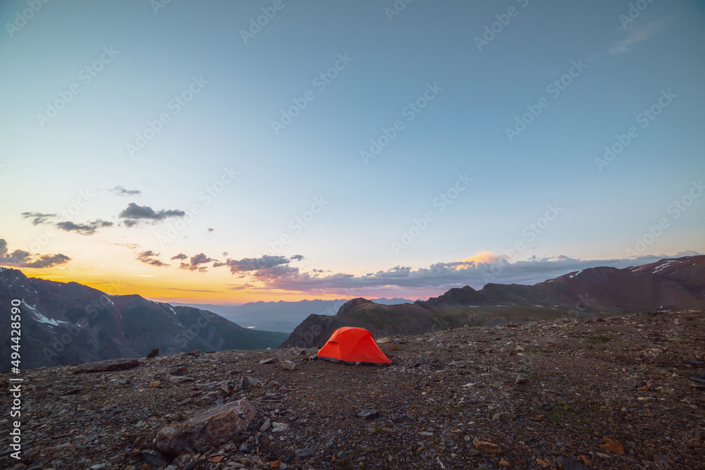 Scenic alpine landscape with tent at very high altitude with view to large mountains in orange dawn sky. Vivid orange tent with awesome view to high mountain range under cloudy sky in sunset colors.