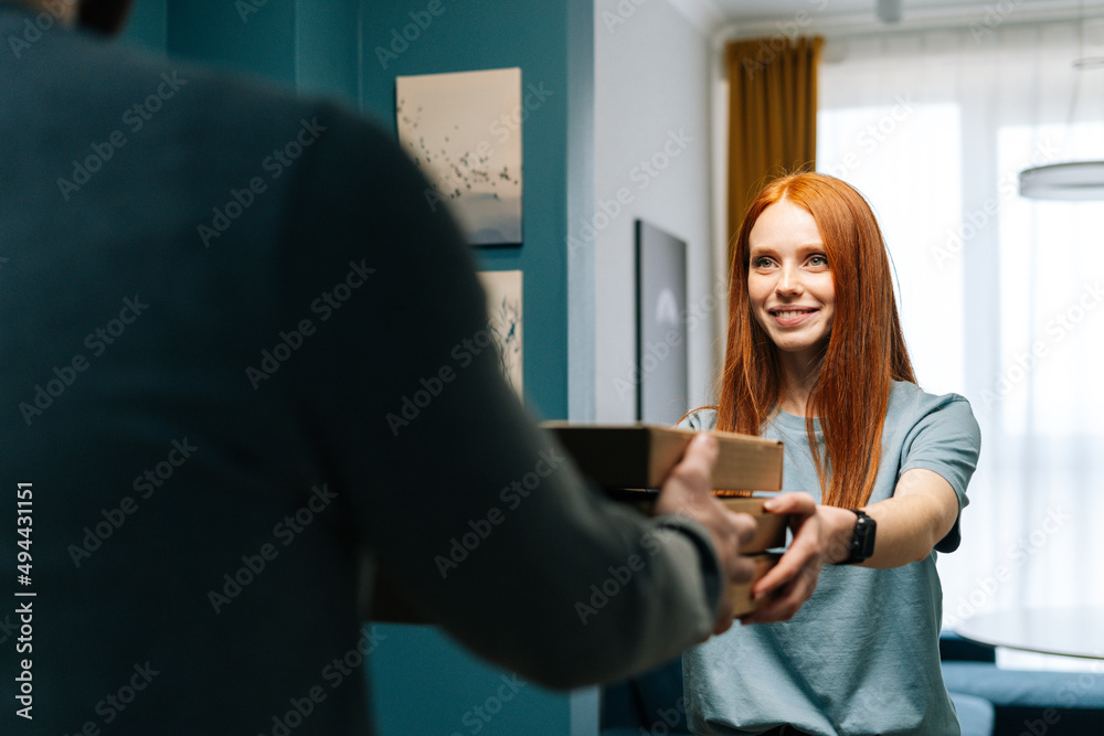 Obraz premium Pretty redhead young woman receiving paper boxes with hot pizza from unrecognizable courier male on doorway at home. Rear view of delivery man delivering boxes with food to female client at apartment.