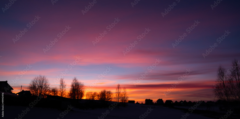 Silhouettes of houses and trees against the bright evening sky. The bright glow of sunset. Pink dawn. Cloudy weather. Scenic natural background. Wallpaper. Scenic view. Twilight in the countryside