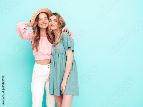 Two young beautiful smiling brunette hipster female in trendy summer dresses. Sexy carefree women posing near blue wall. Positive models having fun. Cheerful and happy. In hats