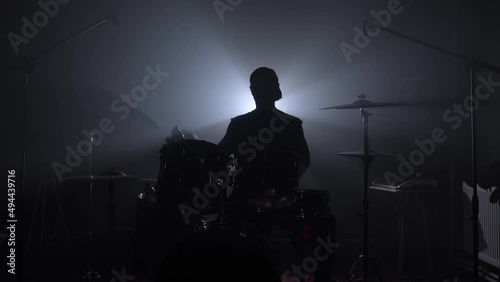 A silhouette of a drummer sits down at a drum kit, twisting his sticks and hitting the cymbal starting to play a recital in a dark, smoke-filled music studio.