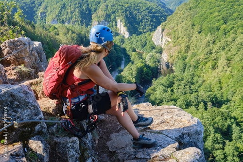 Woman climber with helmet and backpack sits down and relaxes at the end of a via ferrata in Suncuius, Romania, gazing towards Crisul Repede defile covered with green forests, in the morning light. photo