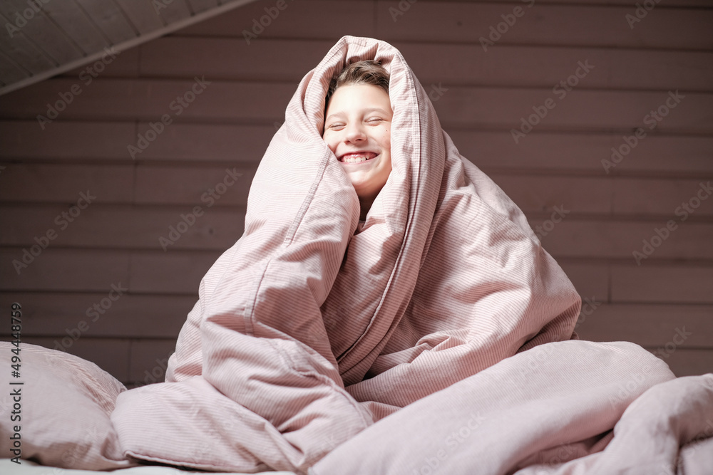Boy teen sitting the bedroom under the pink blanket over head. Smiling