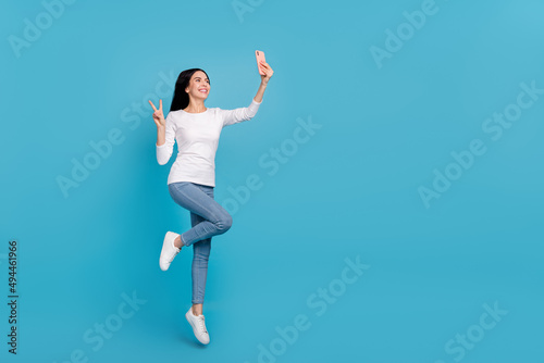 Full length body size view of beautiful trendy girly girl jumping taking selfie showing v-sign isolated on bright blue color background