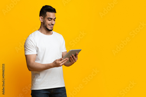 Cheerful arabic guy using digital tablet, yellow studio background