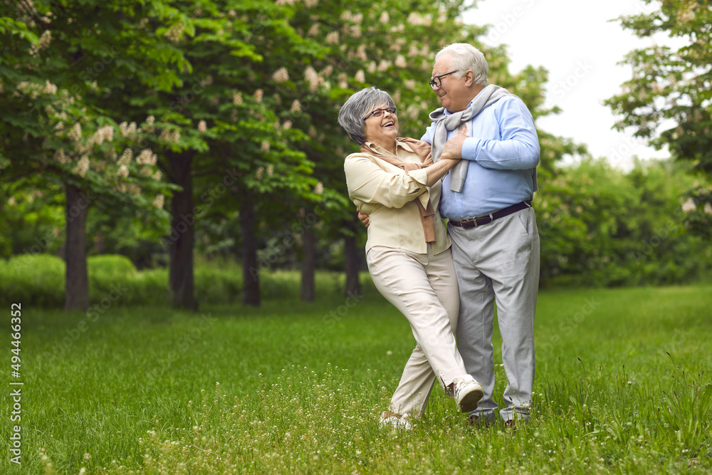 Senior man and woman are dancing on the green grass in the park among the trees. A man holds a woman, wear glasses, and a scarf, happy family life. Walk in the fresh air.