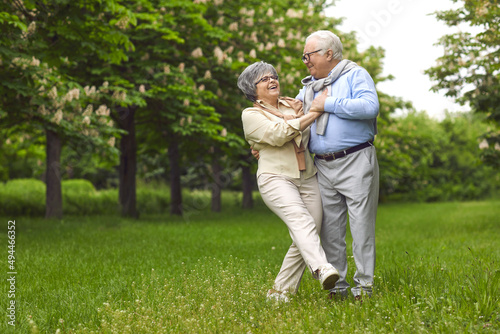 Senior man and woman are dancing on the green grass in the park among the trees. A man holds a woman, wear glasses, and a scarf, happy family life. Walk in the fresh air. © Studio Romantic