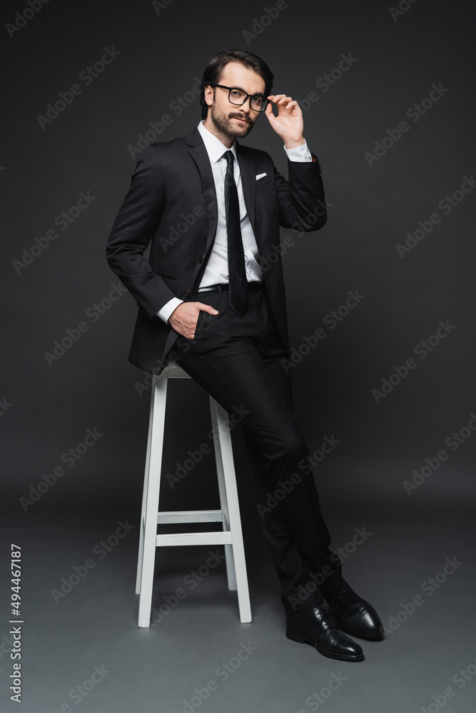 full length of businessman adjusting glasses and leaning on chair while posing with hand in pocket on dark grey. full length of businessman adjusting glasses and leaning on chair while posing with hand in pocket on dark grey.