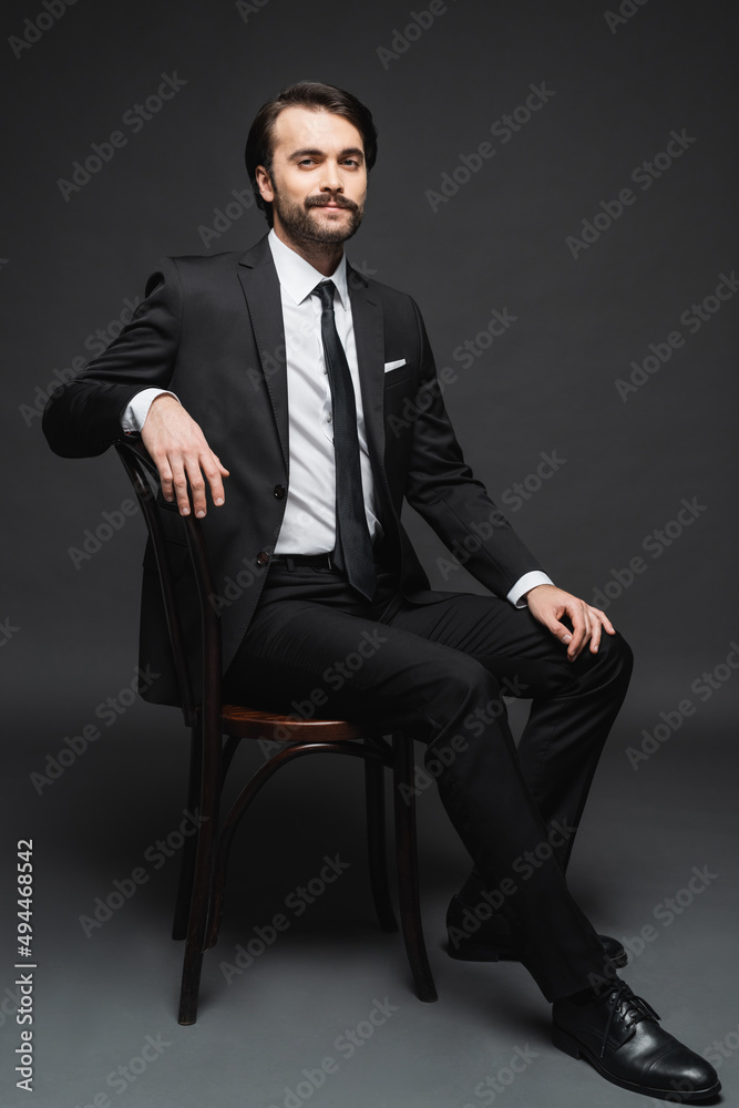 full length of smiling businessman with mustache sitting on wooden chair on dark grey. full length of smiling businessman with mustache sitting on wooden chair on dark grey.