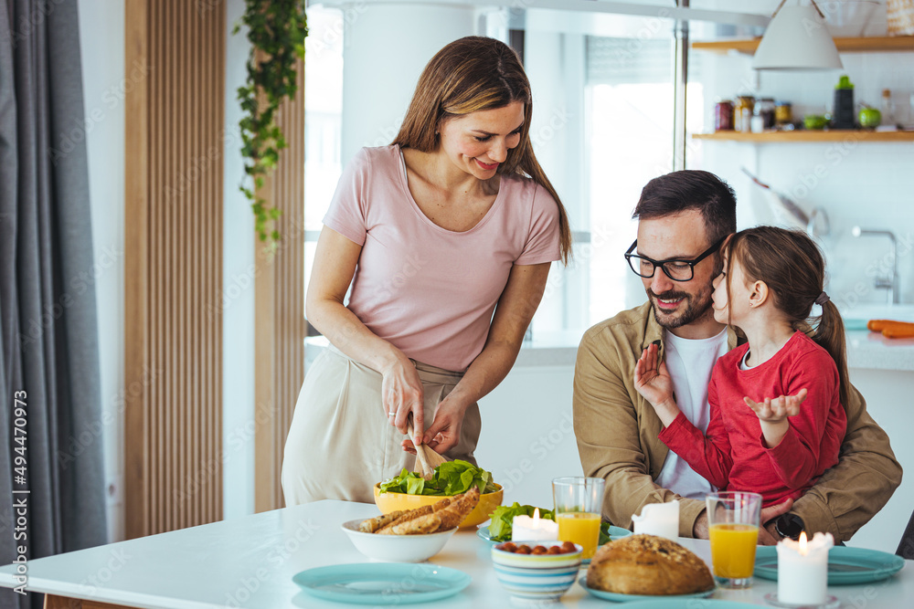 A happy young mother and father with a beautiful daughter cook and have lunch together at home, a smiling mother and little girl have fun preparing healthy food in the kitchen