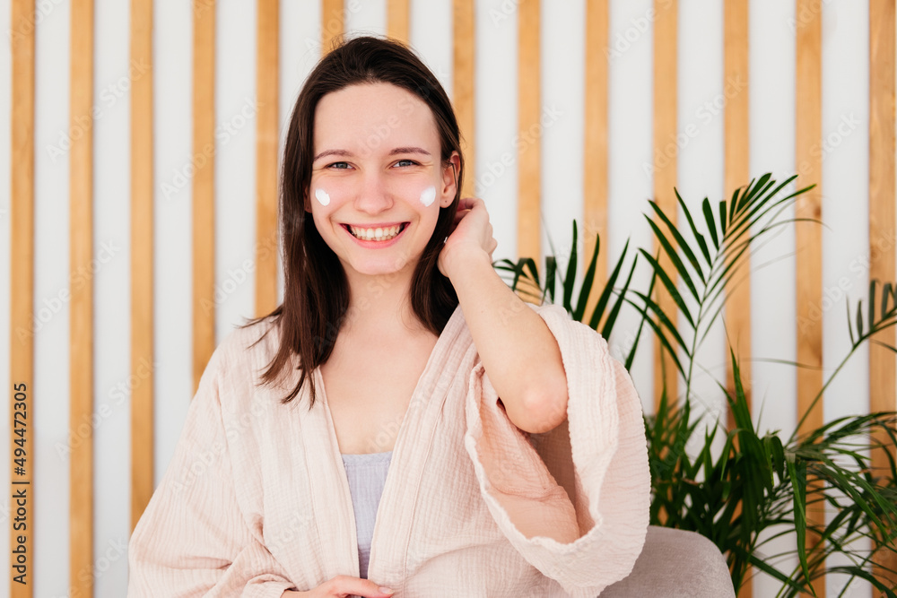 Smiling caucasian young woman applying cream on face at home looking at camera. Morning beauty routine. Skincare concept Smiling caucasian young woman applying cream on face at home looking at camera. Morning beauty routine. Skincare concept