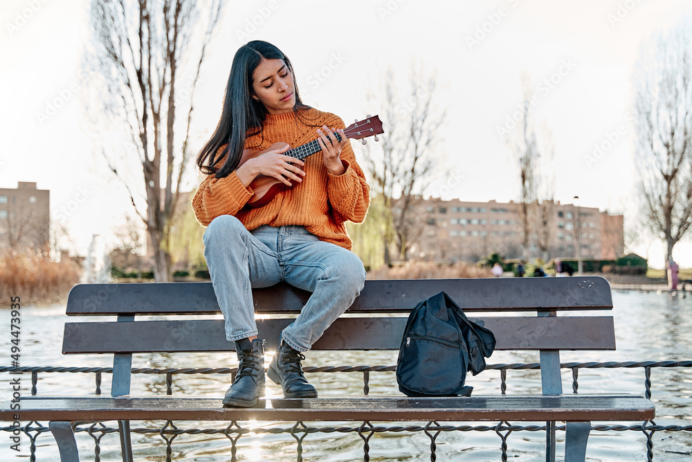 young latina playing ukulele in a city park. woman sitting on a bench practicing with her musical instrument. - obrazy, fototapety, plakaty 