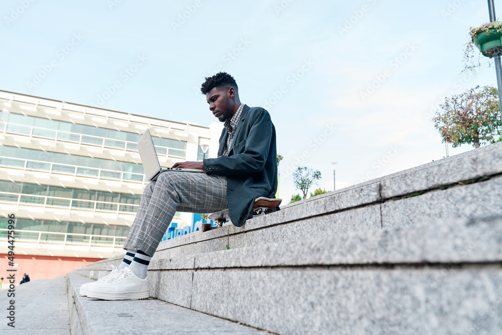 side view of an african american man working with his laptop sitting on a staircase in the city, wearing casual clothes. side view of an african american man working with his laptop sitting on a staircase in the city, wearing casual clothes.