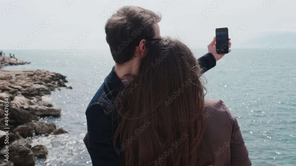 Back view of oving couple of travelers a guy and a girl have a video call conversation on the phone in a picturesque place on the ocean. Young man and woman take a selfie on the beach. High quality 4k Back view of oving couple of travelers a guy and a girl have a video call conversation on the phone in a picturesque place on the ocean. Young man and woman take a selfie on the beach. High quality 4k