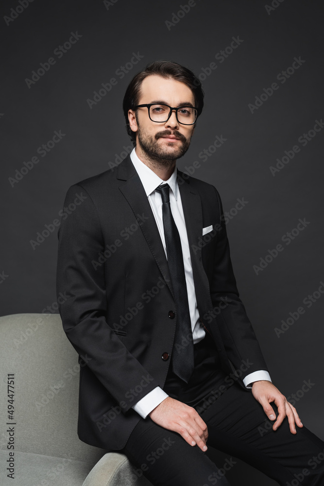 businessman in formal wear and glasses leaning on armchair on dark grey. businessman in formal wear and glasses leaning on armchair on dark grey.