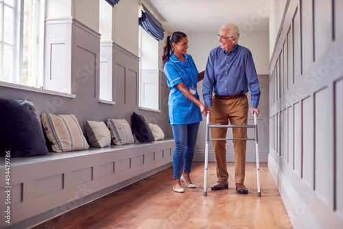 Senior Man At Home Using Walking Frame Being Helped By Female Care Worker In Uniform photo