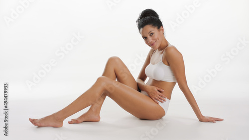 Long shot of African American slim gorgeous woman in white underwear sitting on the floor, looking at camera smiling on white background | After leg shaving concept