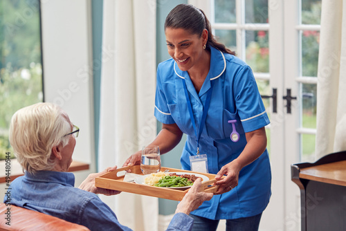 Female Care Worker In Uniform Bringing Meal On Tray To Senior Man Sitting In Lounge At Home  photo