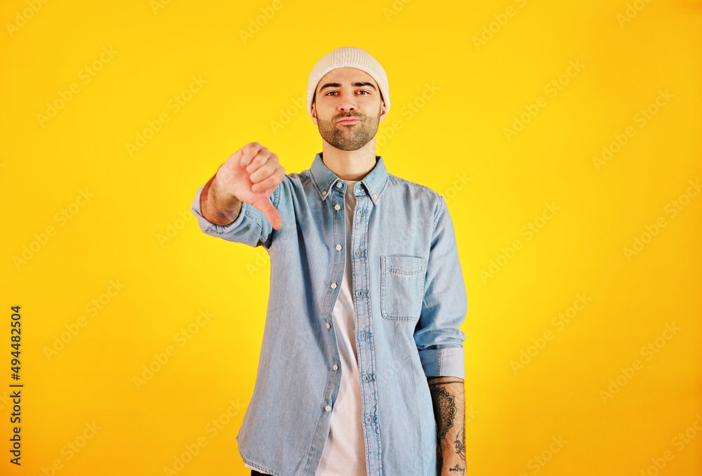 Showing dislike. Studio shot of smiliing handsome man in jeans and white hat on yellow background. Tatoo and beard. Showing dislike. Studio shot of smiliing handsome man in jeans and white hat on yellow background. Tatoo and beard.