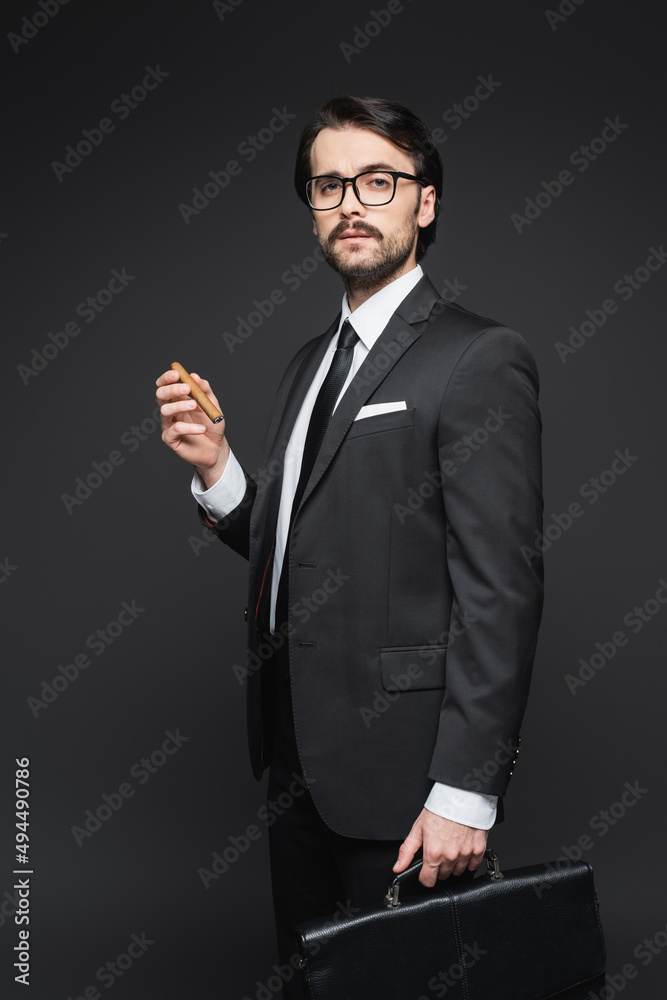 businessman in suit and glasses holding cigar and leather briefcase on dark grey. businessman in suit and glasses holding cigar and leather briefcase on dark grey.