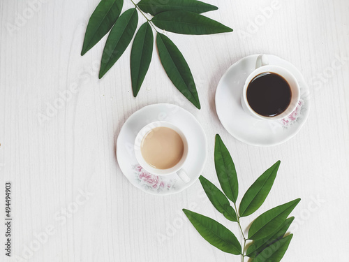  white cup with black coffee and one with coffee and milk on white wooden background with leaves and space for text