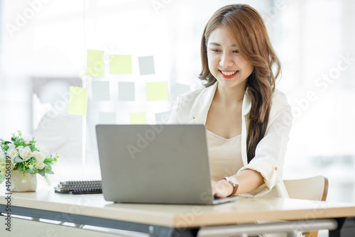 Portrait of Asian young businesswoman with laptop writes on a document at her office Asian girl working at a office space with a laptop computer. 