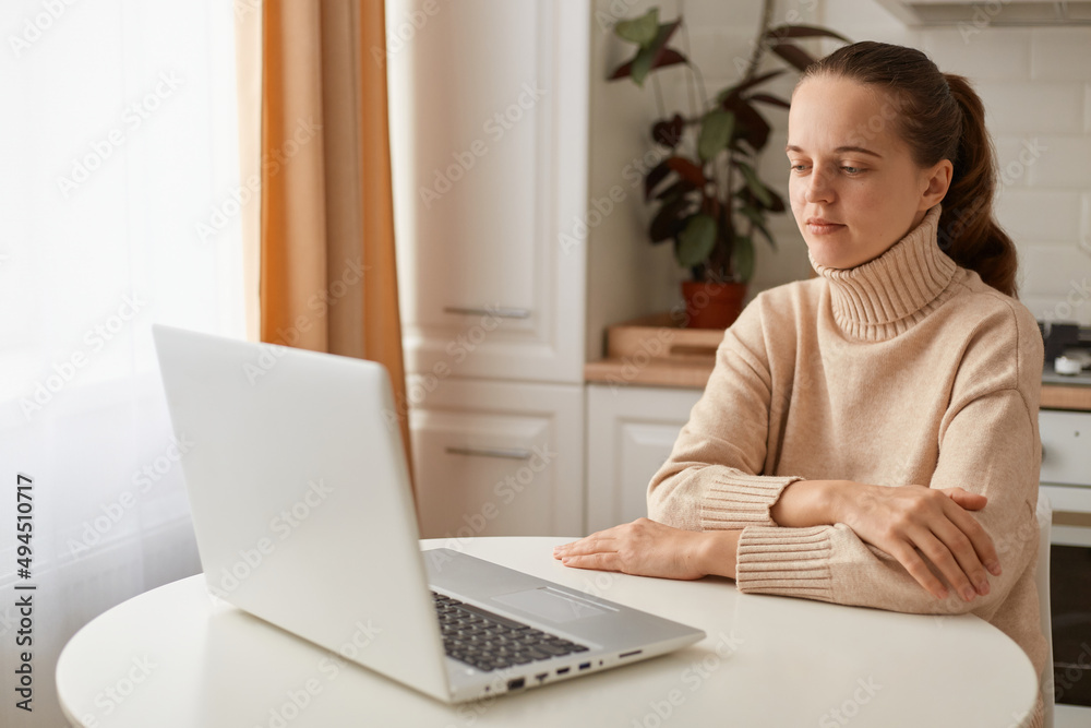 Indoor shot of attractive young adult woman with dark hair wearing beige sweater sitting in kitchen and working on laptop or watching video, having video call, listening online lecture. Indoor shot of attractive young adult woman with dark hair wearing beige sweater sitting in kitchen and working on laptop or watching video, having video call, listening online lecture.