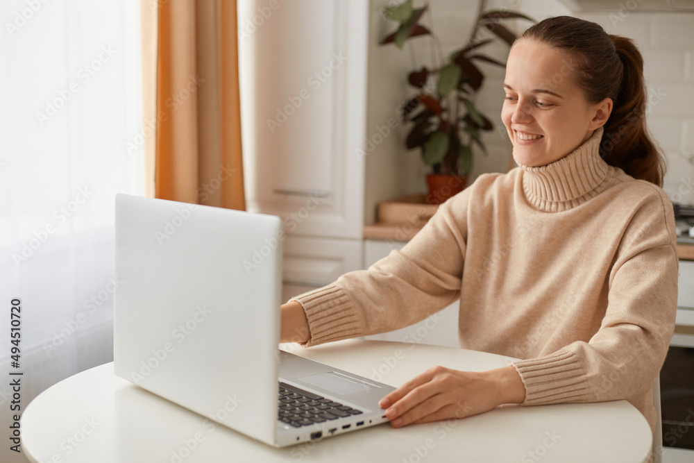 Smiling satisfied woman with dark hair wearing beige sweater sitting in kitchen and working on laptop, looking at display with happy positive facial expression. Smiling satisfied woman with dark hair wearing beige sweater sitting in kitchen and working on laptop, looking at display with happy positive facial expression.