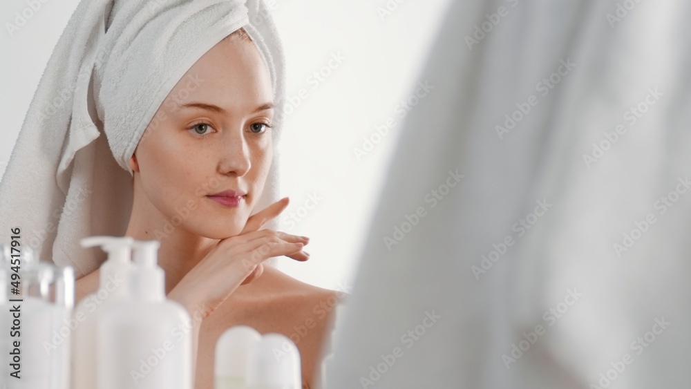 Gorgeous slim Caucasian young woman with a hair towel touches her face and looks in the bathroom mirror | Face care concept Gorgeous slim Caucasian young woman with a hair towel touches her face and looks in the bathroom mirror | Face care concept