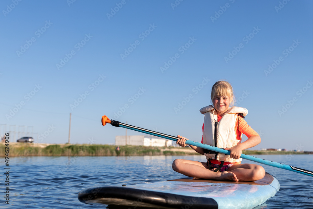 Cute little caucasian blond child girl enjoy having fun sitting on sup board surfing at freshwater pond lake or river hot sunny day. Summer outdoors child healthy sport recreation activities