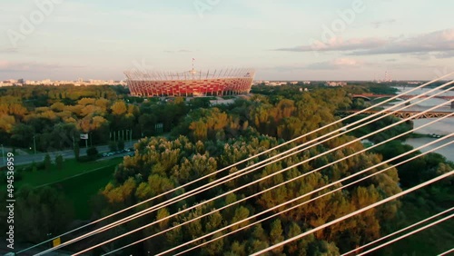 Aerial view of National Stadium known as Stadion Narodowy in Warsaw, Poland. Roof Football arena with red and white facade. Swietokrzyski Bridge and Vistula River. 4K zoom out shot photo