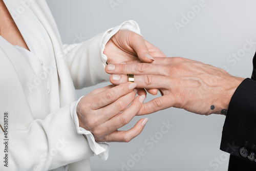 Cropped view of mature woman wearing wedding ring on groom isolated on grey.
