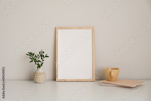 Vertical picture frame mockup on white table. Vase with green branch, mug of tea, paper notebook. White wall background. Nordic style living room interior design