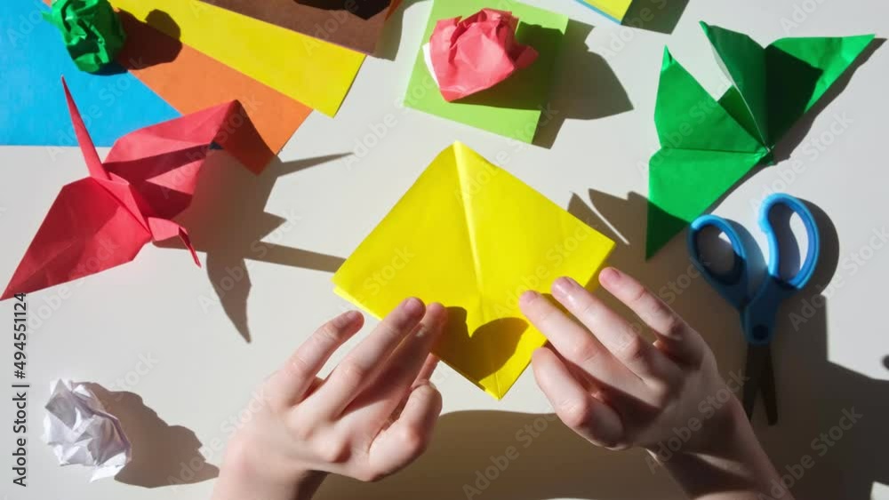 Children's hands do origami from colored paper on white background. Origami paper. DIY. Various school supplies. Concept of children's creativity, back to school.	
