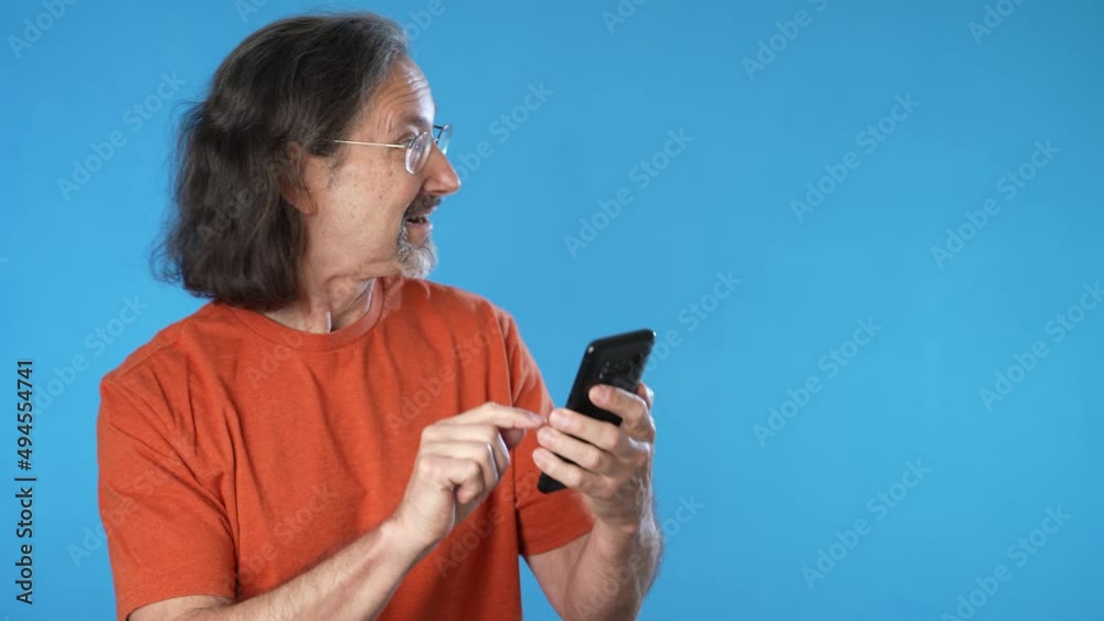 Surprised joyful mature man hold mobile cell phone typing say wow success points to product sale, isolated on solid blue background studio copy space. Surprised joyful mature man hold mobile cell phone typing say wow success points to product sale, isolated on solid blue background studio copy space.