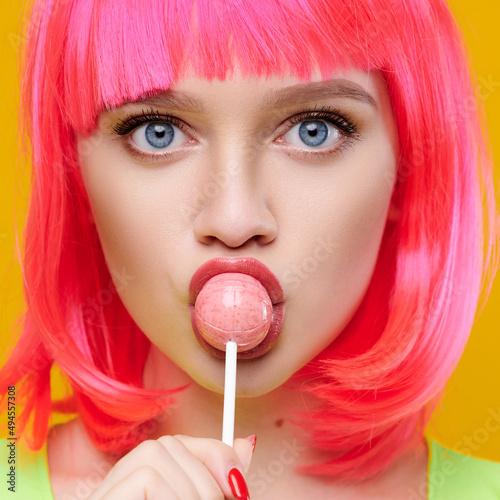 Freaky attractive hipster woman in pink wig eat lick lollipop on bright yellow background