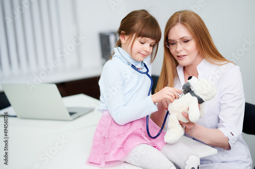 Caucasian female doctor wear face mask show little girl examining bear toy by stethoscope indoor hospital