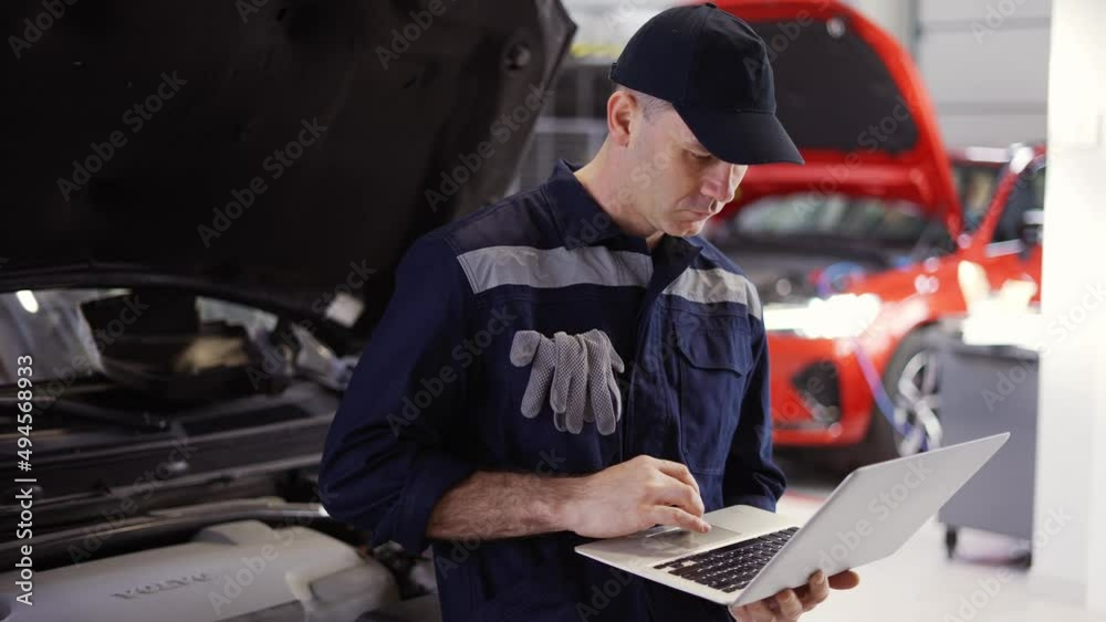 Car mechanic working on laptop in auto repair service, lean on a car Car mechanic working on laptop in auto repair service, lean on a car