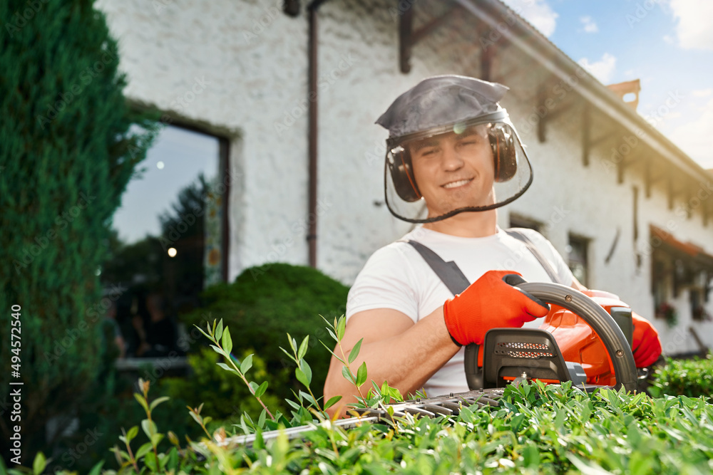 Caucasian man wearing protective mask and gloves pruning hedge with electric trimmer during summer days. Gardener working with modern equipment on fresh air. Caucasian man wearing protective mask and gloves pruning hedge with electric trimmer during summer days. Gardener working with modern equipment on fresh air.