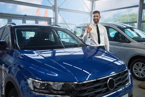 Smiling caucasian businessman gesturing sign OK with finger while standing near luxury modern car. Happy bearded person buying new transport at auto salon. © serhiibobyk