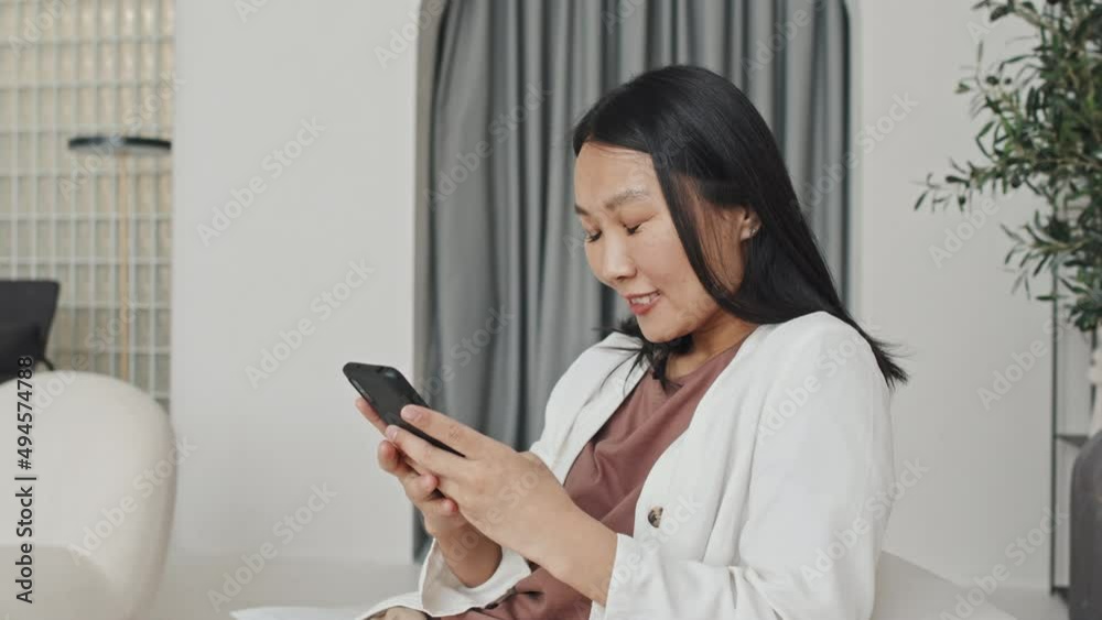 Medium of young cheerful Asian woman typing on smartphone, sitting in house with modern minimalist interior design at daytime, smiling Medium of young cheerful Asian woman typing on smartphone, sitting in house with modern minimalist interior design at daytime, smiling