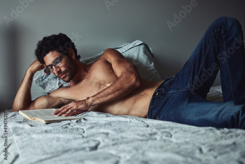 He enjoys reading novels over the weekend. Cropped shot of a handsome young shirtless man reading a book on his bed at home.