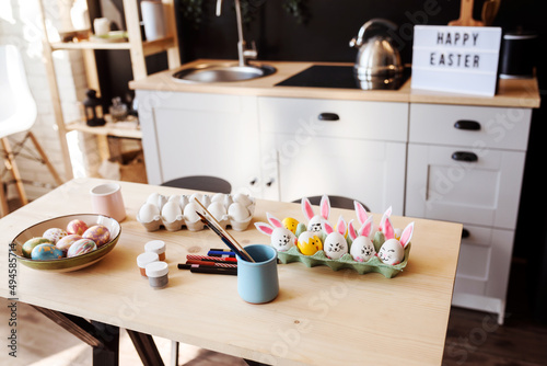 Easter holiday preparations, dyed Easter eggs, paints and brushes on the table in the kitchen. Happy Easter congratulation written on the table in the background. 