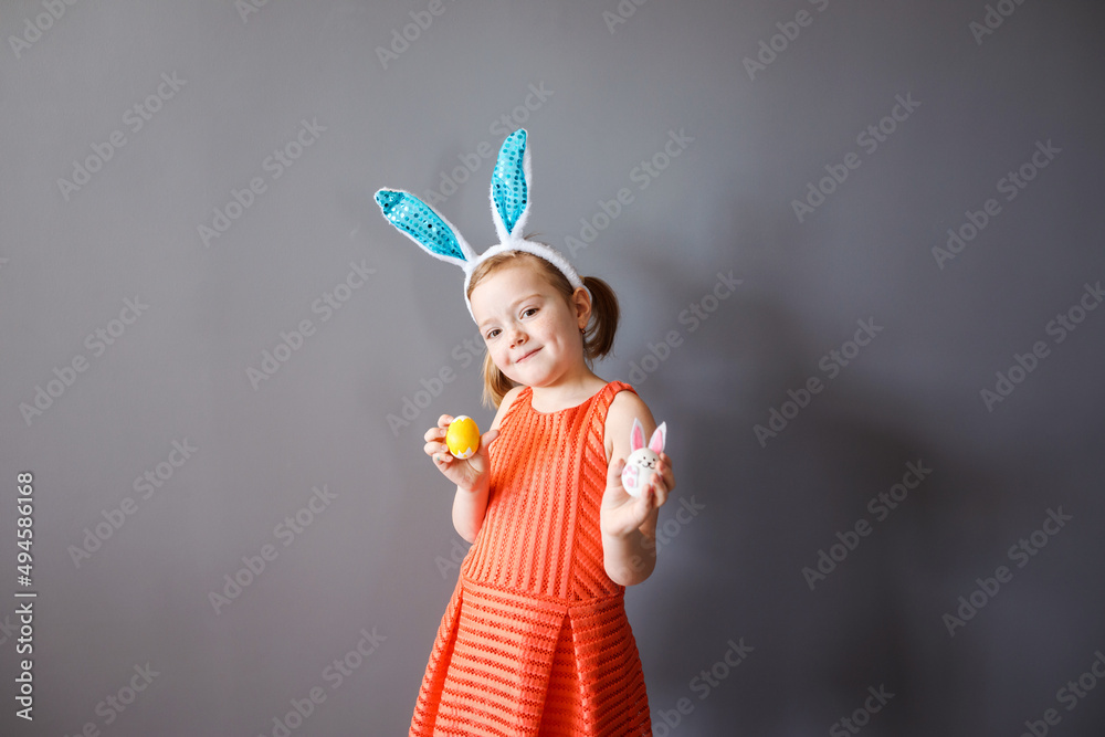 Funny little girl in bunny ears headband with Easter eggs in her hands in front of gray background