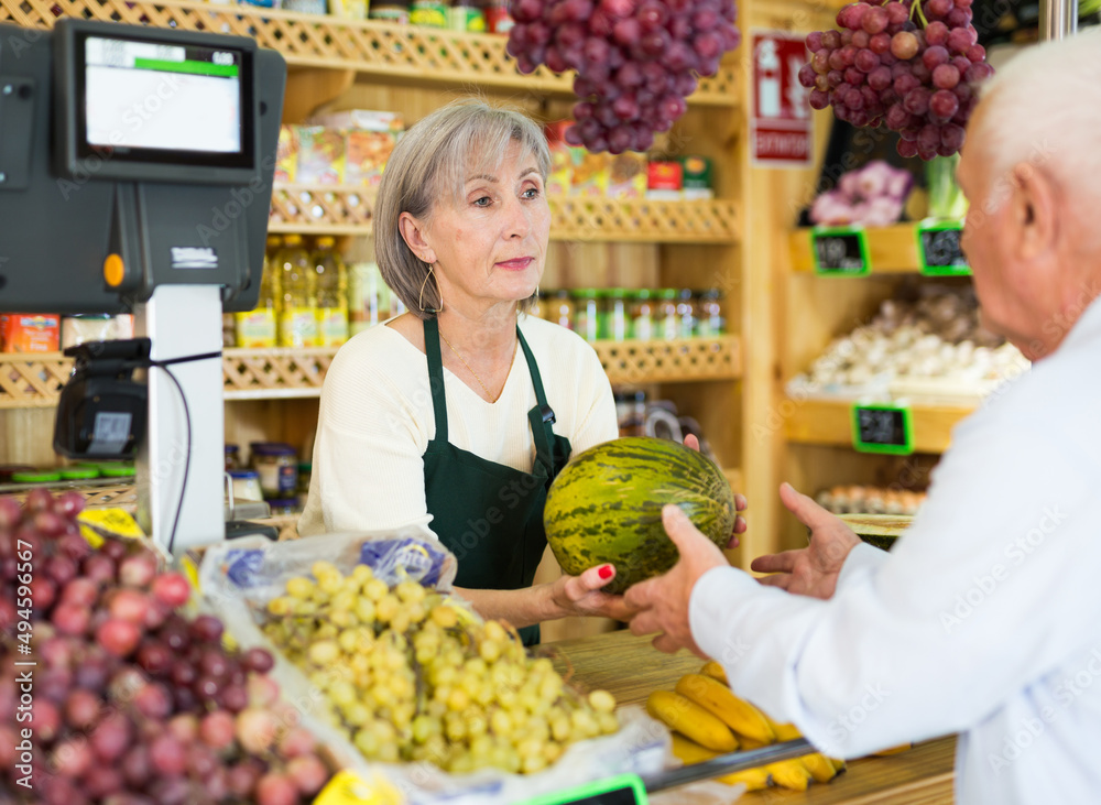 Woman supermarket employee hands watermelon to an elderly man after paying for purchase at the checkout