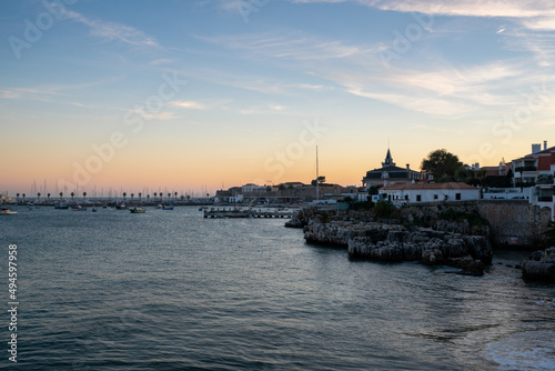 Rainha beach in Cascais at sunset with sail boats on the sea and houses on the coast, in Portugal © Luis