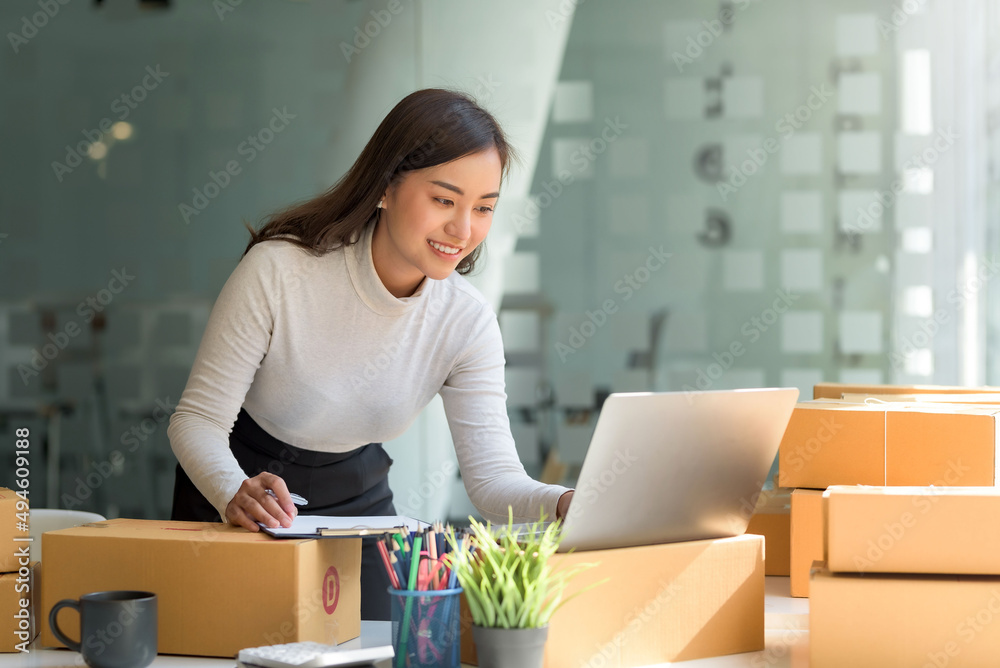 Happy young Asian business owner woman prepare parcel box and check online orders of product for deliver to customer on laptop computer. Shopping Online concept. - obrazy, fototapety, plakaty 
