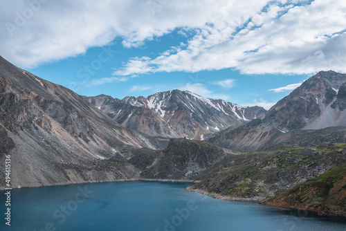 Deep mountain lake of phantom blue color among sharp rocks and high mountains in changeable weather. Wonderful dramatic view to deep blue mountain lake and large sunlit rocky cliff under cloudy sky. © Daniil